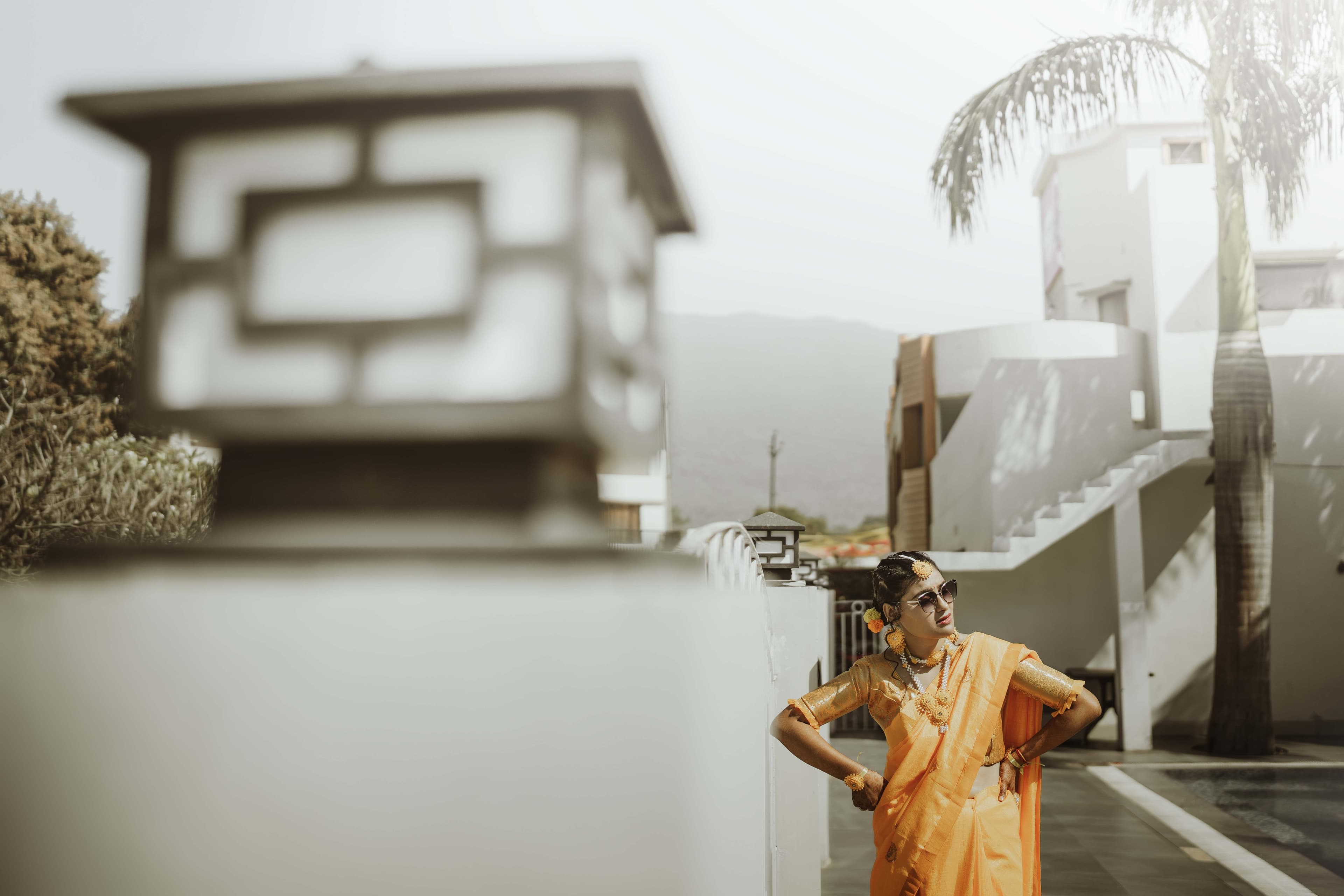 A woman in an orange sari stands gracefully beside a building, captured in a candid moment of beauty and culture